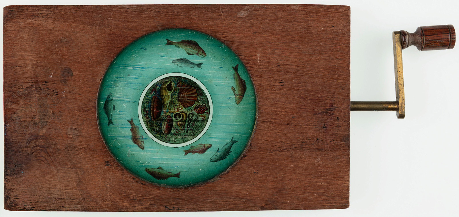 Automated timber and brass framed circular glass lantern slide featuring fish and other marine creatures. Timber and brass handle operates secondary plate allowing the fish to move.