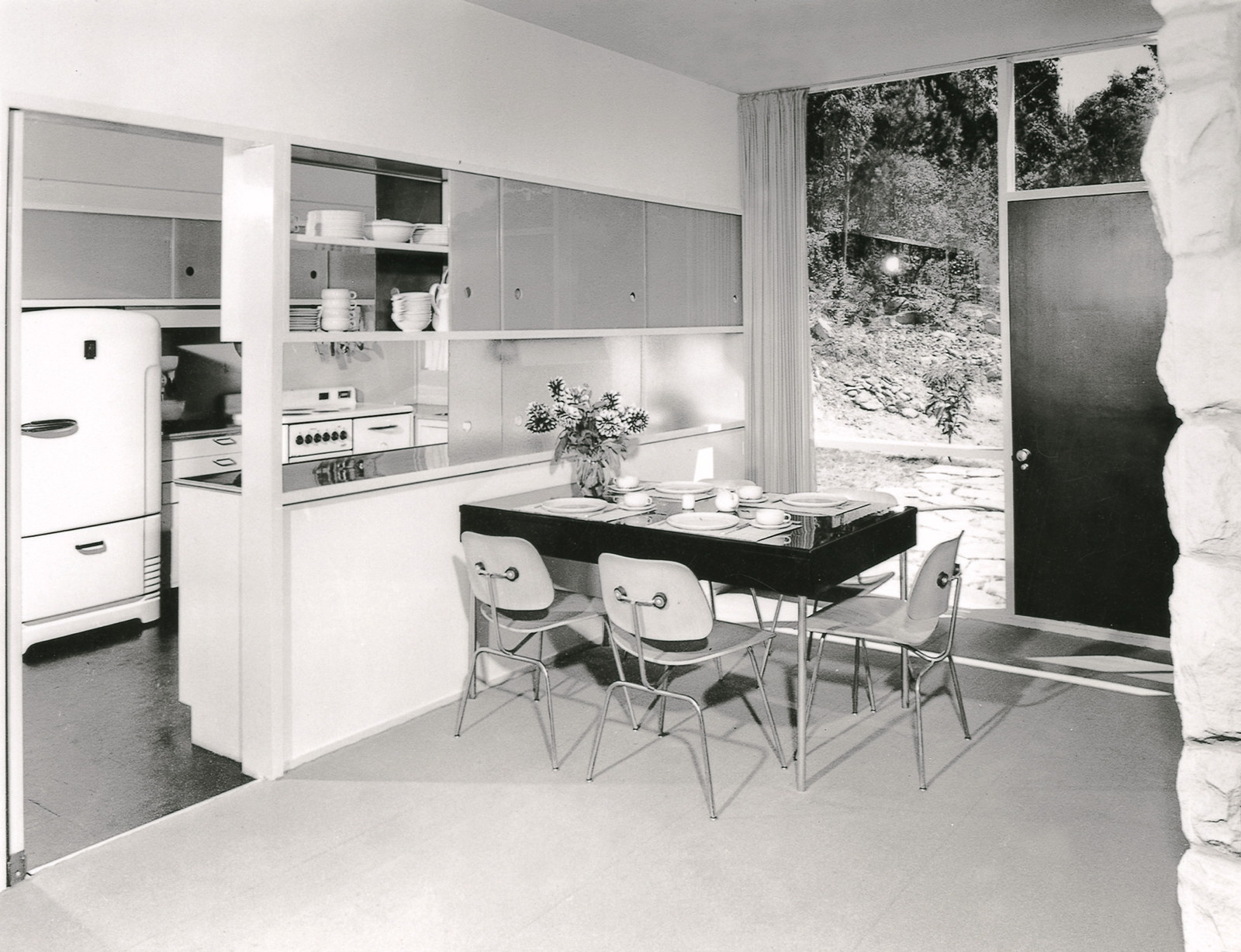 Black and white photo of dining room table and chairs with window and kitchen in background.