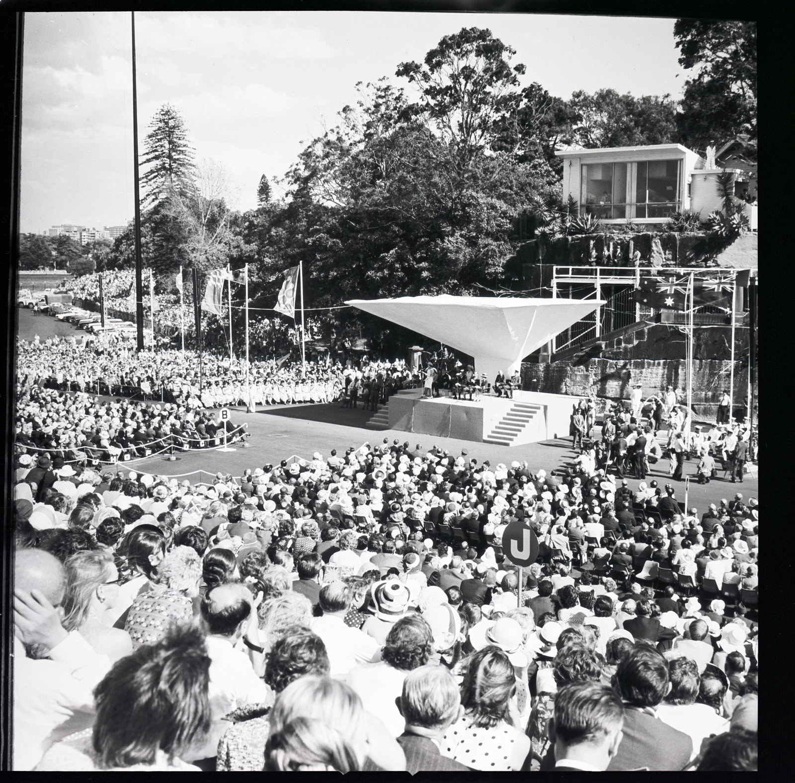 HM the Queen delivering Her speech at the opening of the Opera House. The official party seated on a dais with inverted roof, set up in the forecourt before the Tarpeian Steps, with a temporary works viewing pavilion behind. October 20th 1973.