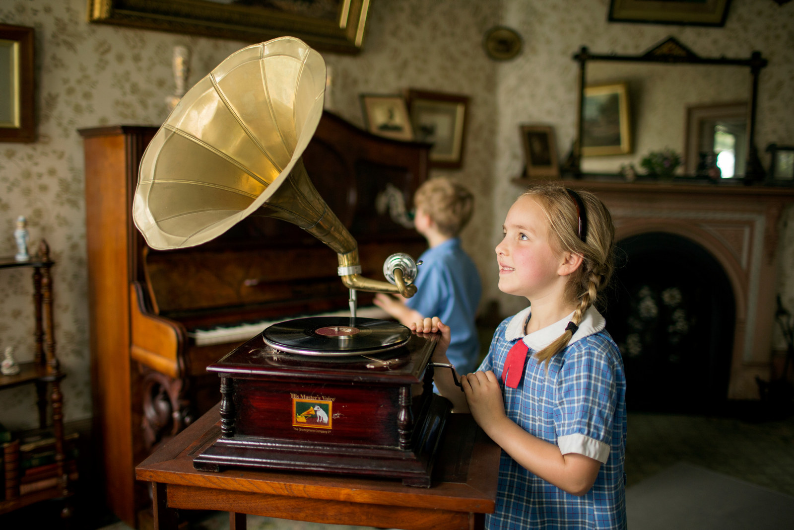 A girl looking at a gramophone with a boy sitting at the piano in the parlour at Meroogal House