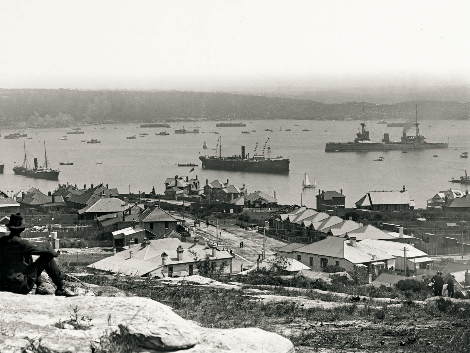 Black and white photograph showing view of harbour with ships with rocks in foreground.