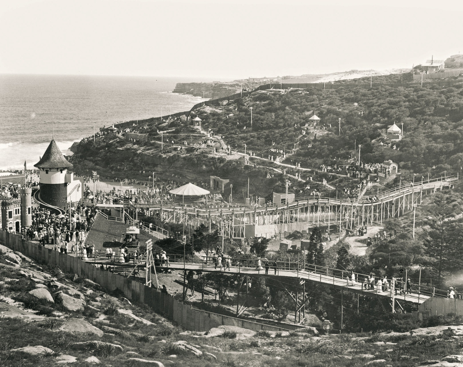 View of the roughed headlands and steep hills leading to Tamarama Beach crowded with people and light with timber walkways and amusement park structures.