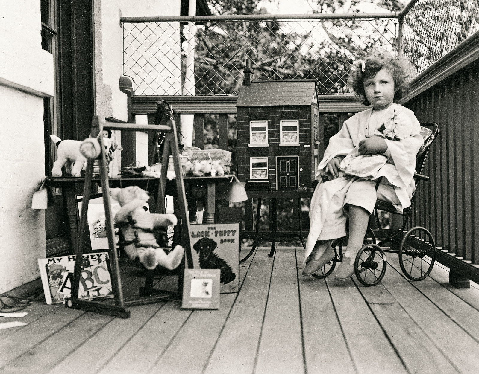 Young girl sits on a chair with wheels on a verandah. She is surrounded by toys including a dolls house and fluffy toy elephant. She holds a teddy bear.
