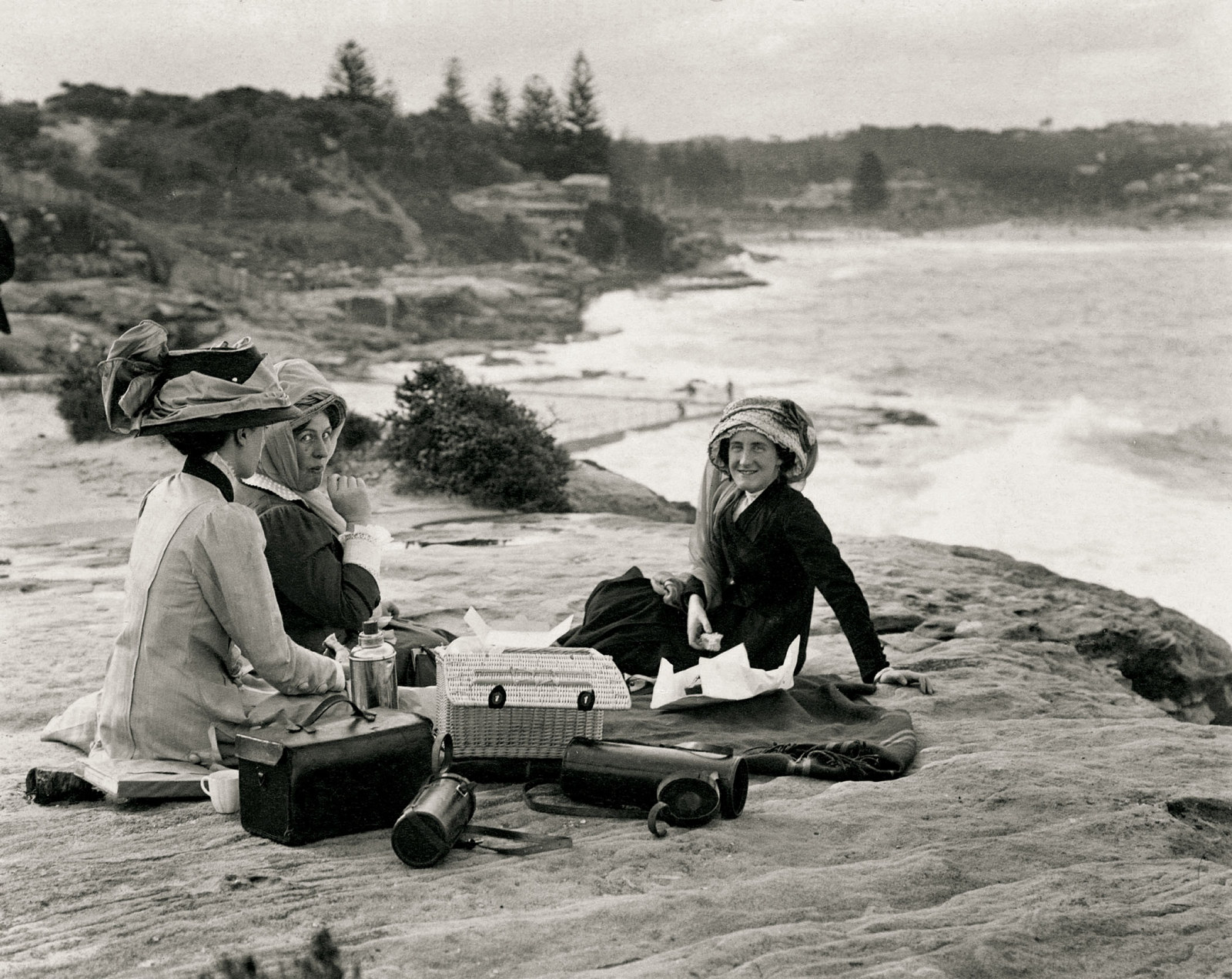 Black and white photograph of group sitting on sand. The women are wearing elaborate motoring hats with scarves.