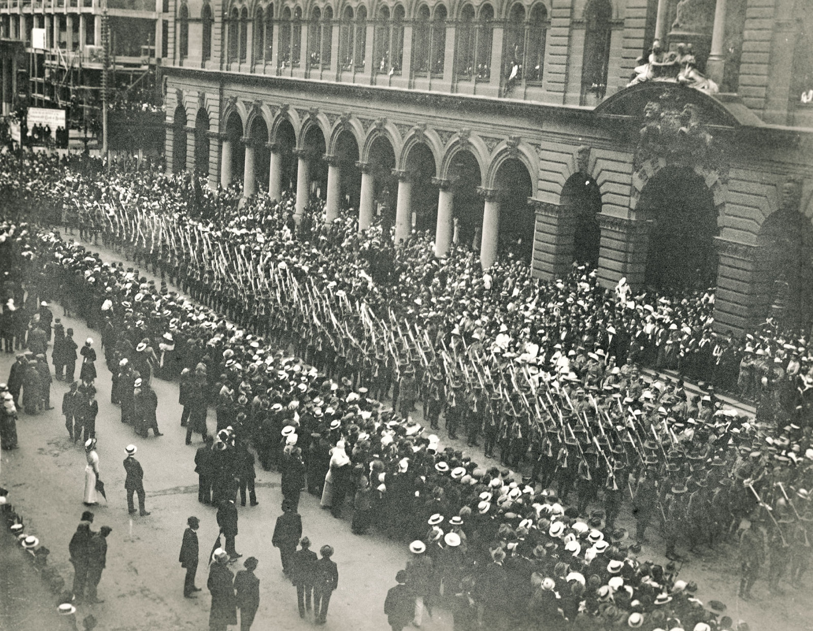View of strret lined with crowds and troops marching down the centre of the street.