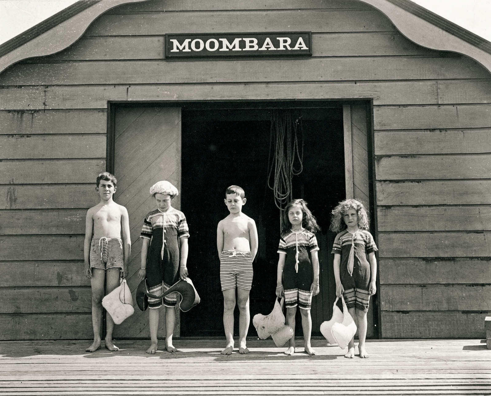 Row of five children stand in front of a timber boatshed. They wear knitted swimming costumes and carry life vests.