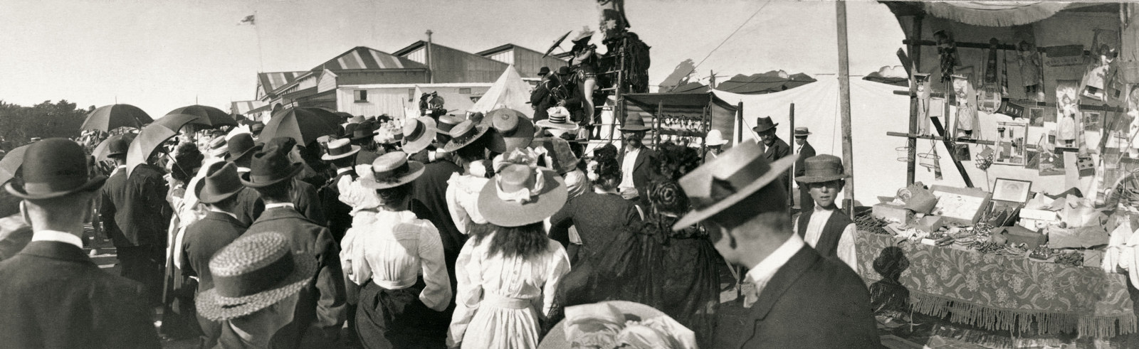 a panoramic image of crowds at a side show. Young girl with long hair and hat is in the foreground. An unclear attraction is on a stage is in the background.