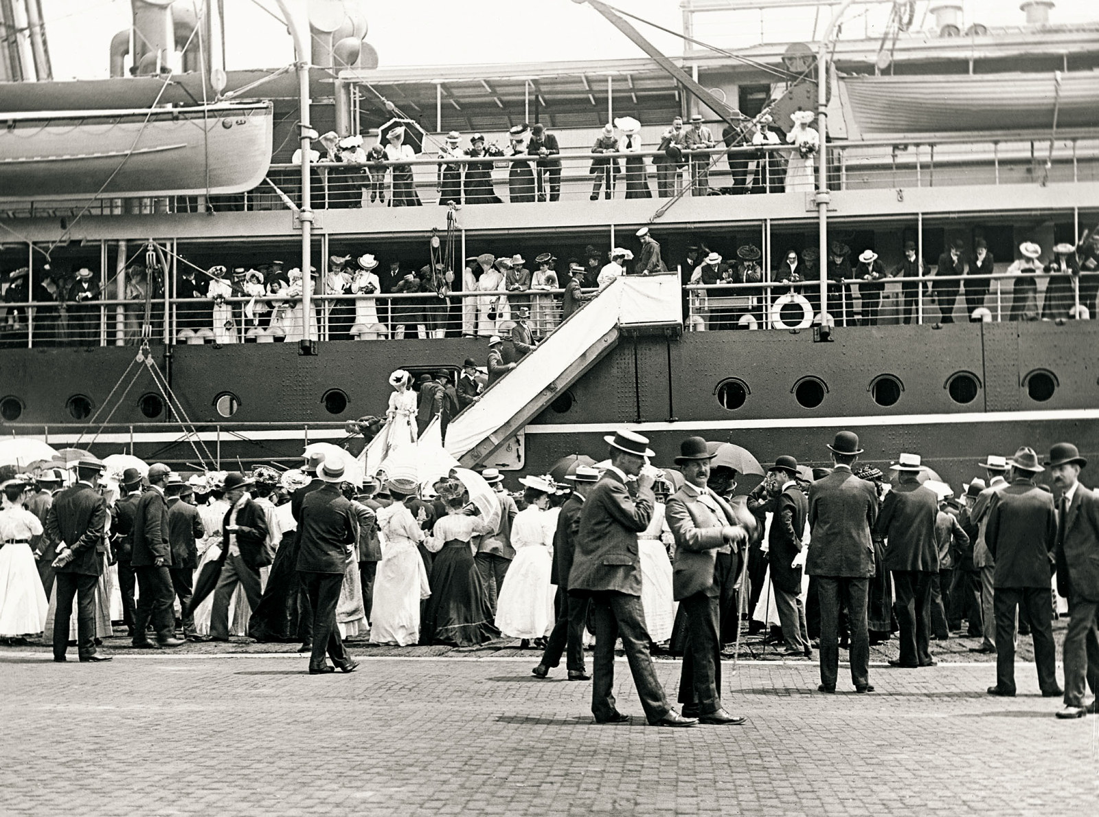 A crowd farewells a large ship at a dock. Two men walk in the foreground wearing suits, waistcoats and hats.