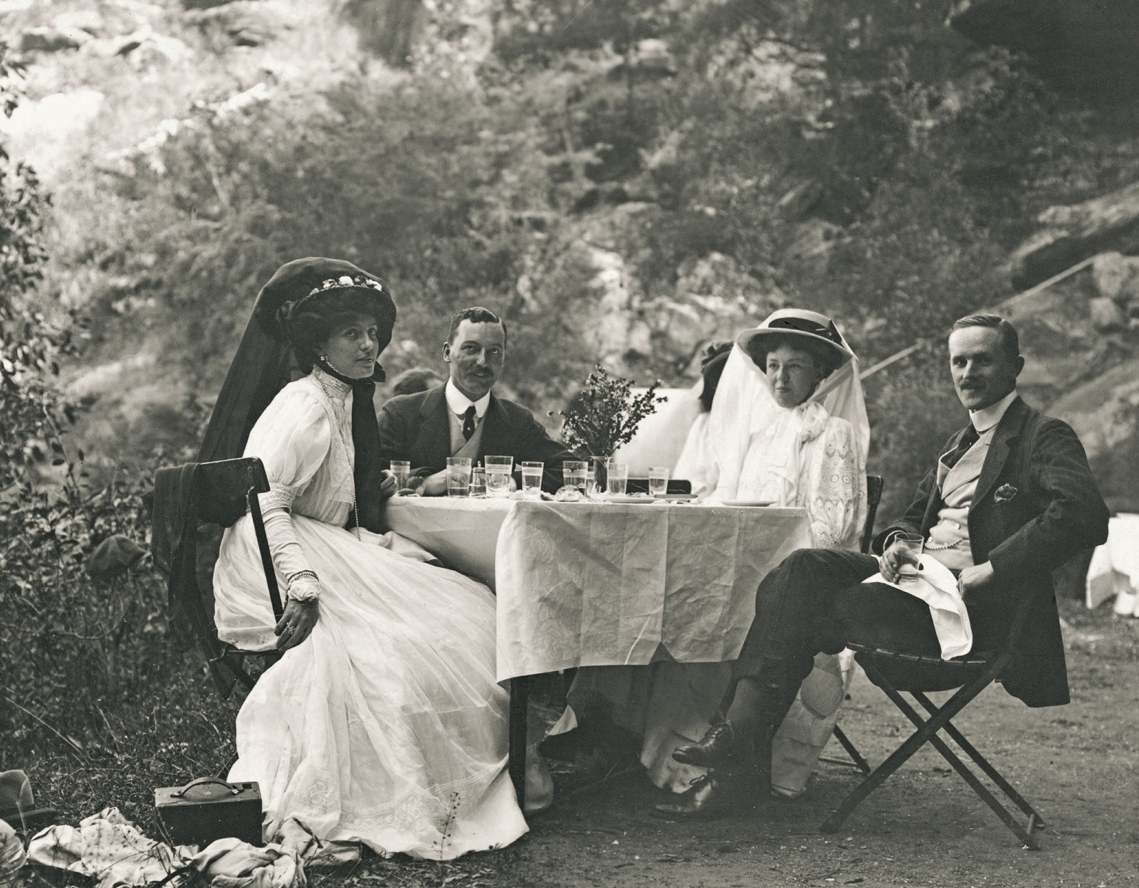 Four people sit outside gathered around a small table with white table cloth, surrounded by trees.