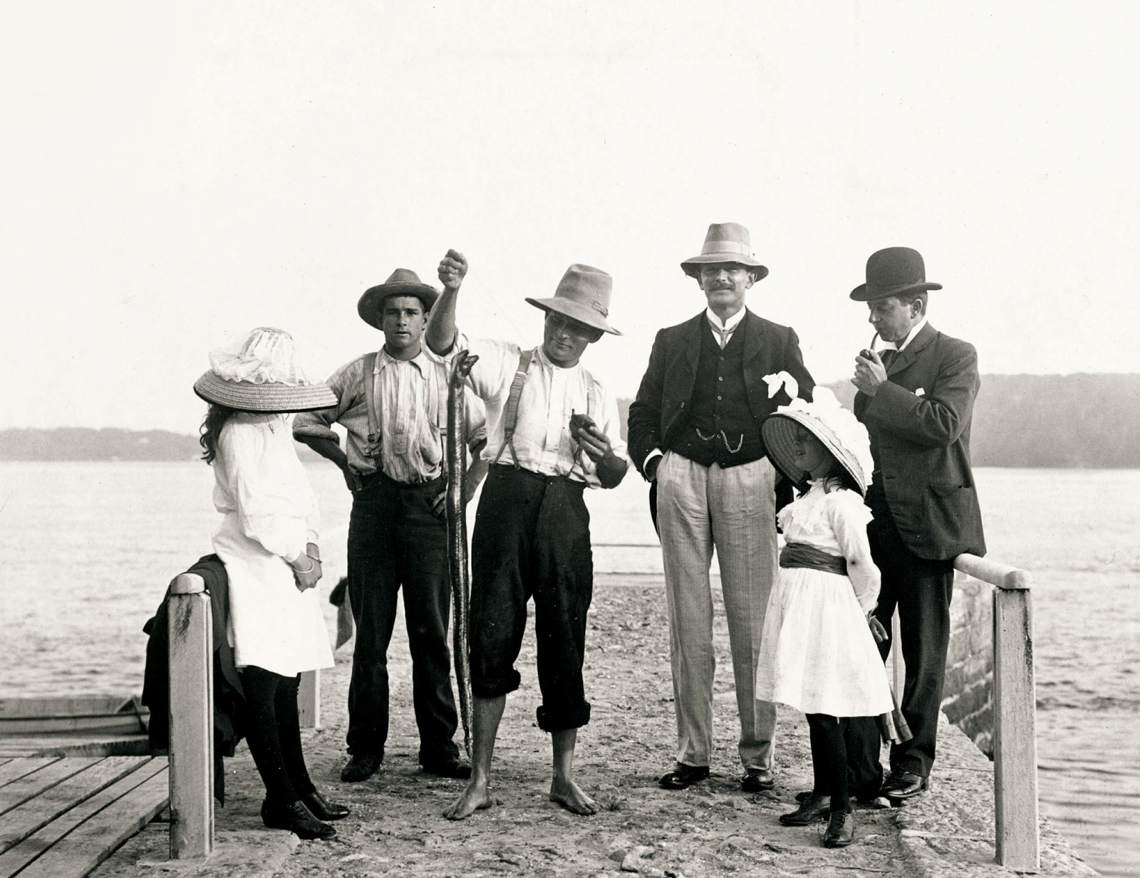 Group of children and adults gather on a jetty. Man in centre holds large eel caught on a fishing hook.