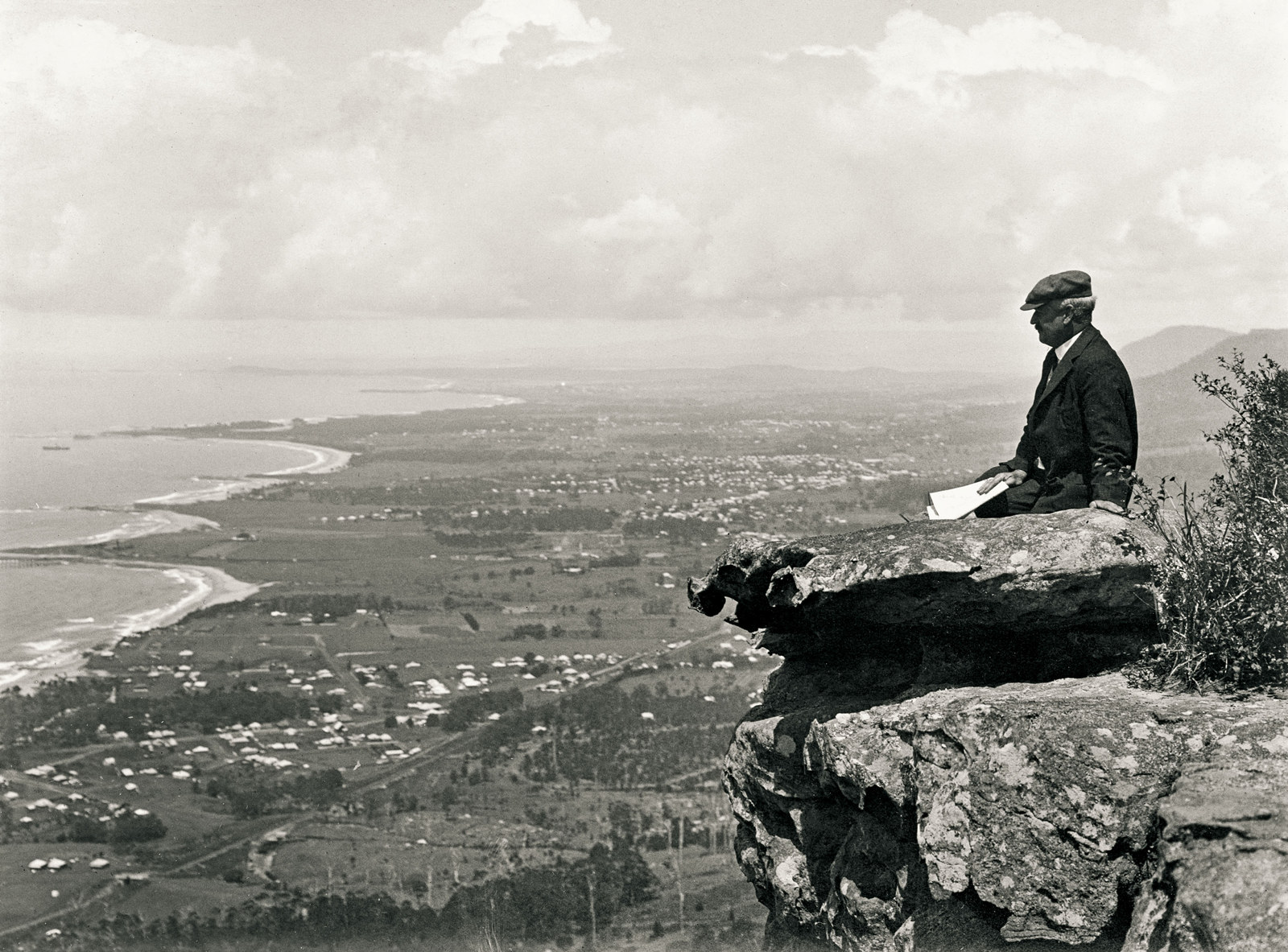 Man sits on a rock at the edge of an escarpment with a dramatic view showing a sweeping coastline, fields and dotted with houses.